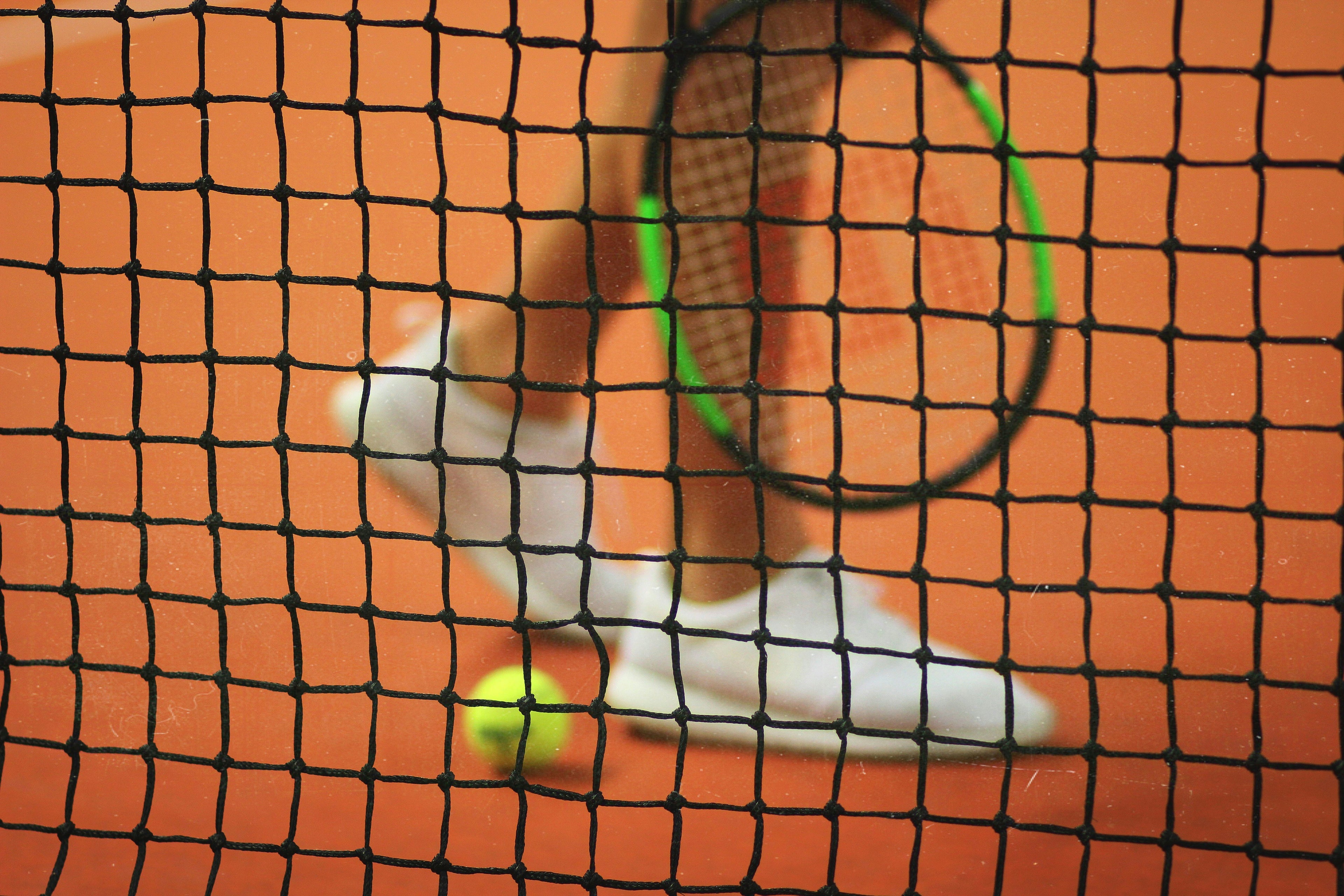 Blurred view through a tennis net capturing dynamic motion of a player with tennis shoes and racket on a clay court, evoking active lifestyle energy.