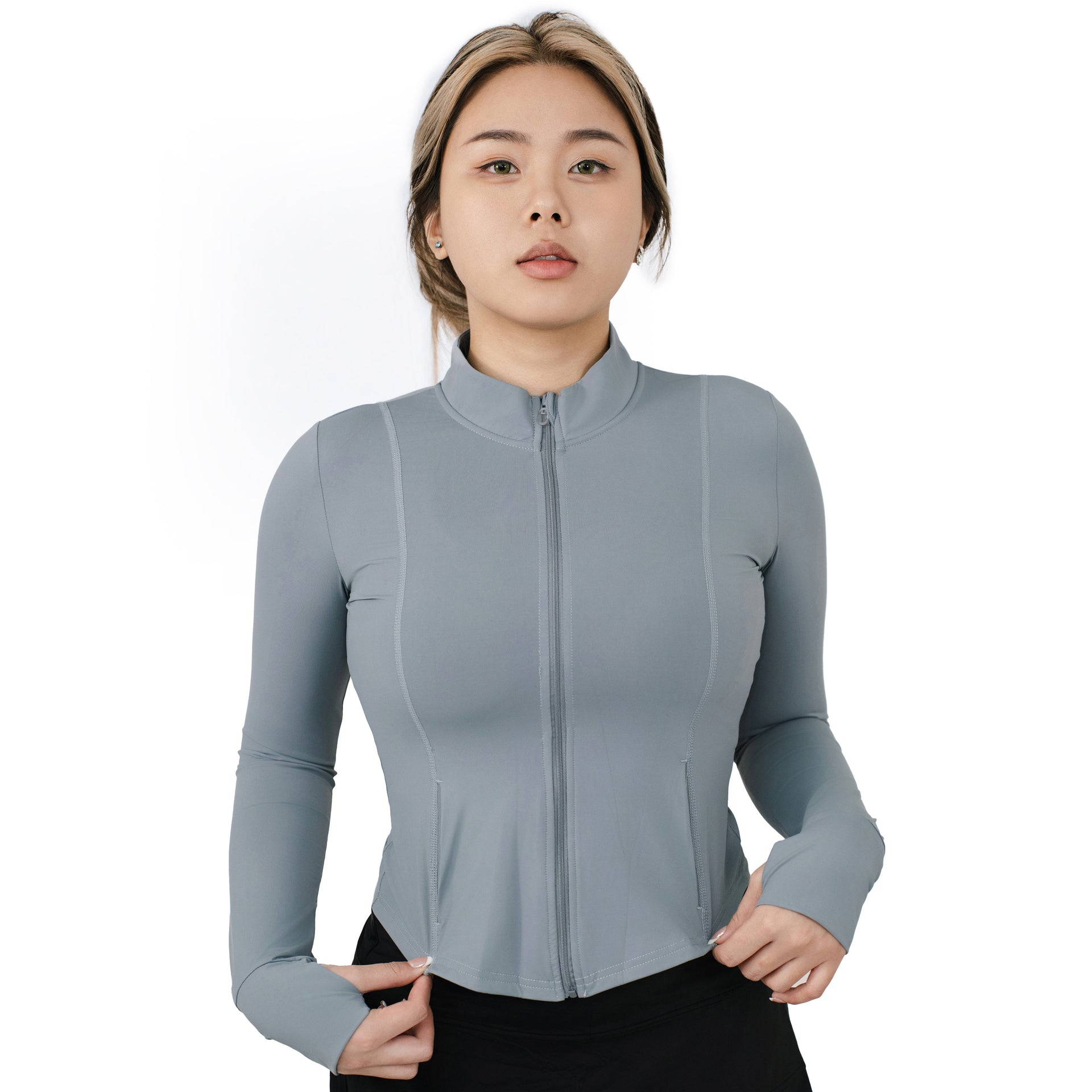 The model wearing VLIZ ash gray full-zip slim fit long sleeve top with mock neck and thumbhole cuffs, facing forward with hands pulling slightly at the hem, ideally for tennis and outdoor workouts.
