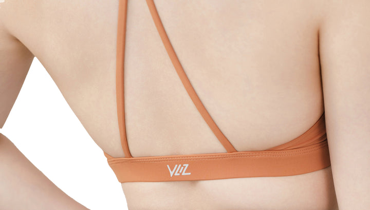 Close-up of the VLIZ SculptFlex Cross-Back Sports Bra in Caramel Orange, showcasing the minimalist criss-cross back strap design and soft elastic underband with logo detail.
