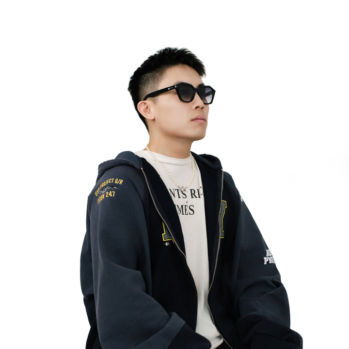 Male model wearing VLIZ Altara sunglasses with a black square-oval frame and grey lenses, styled with a hoodie and layered necklace, emphasizing the unisex streetwear-inspired design and UV400 lens protection.