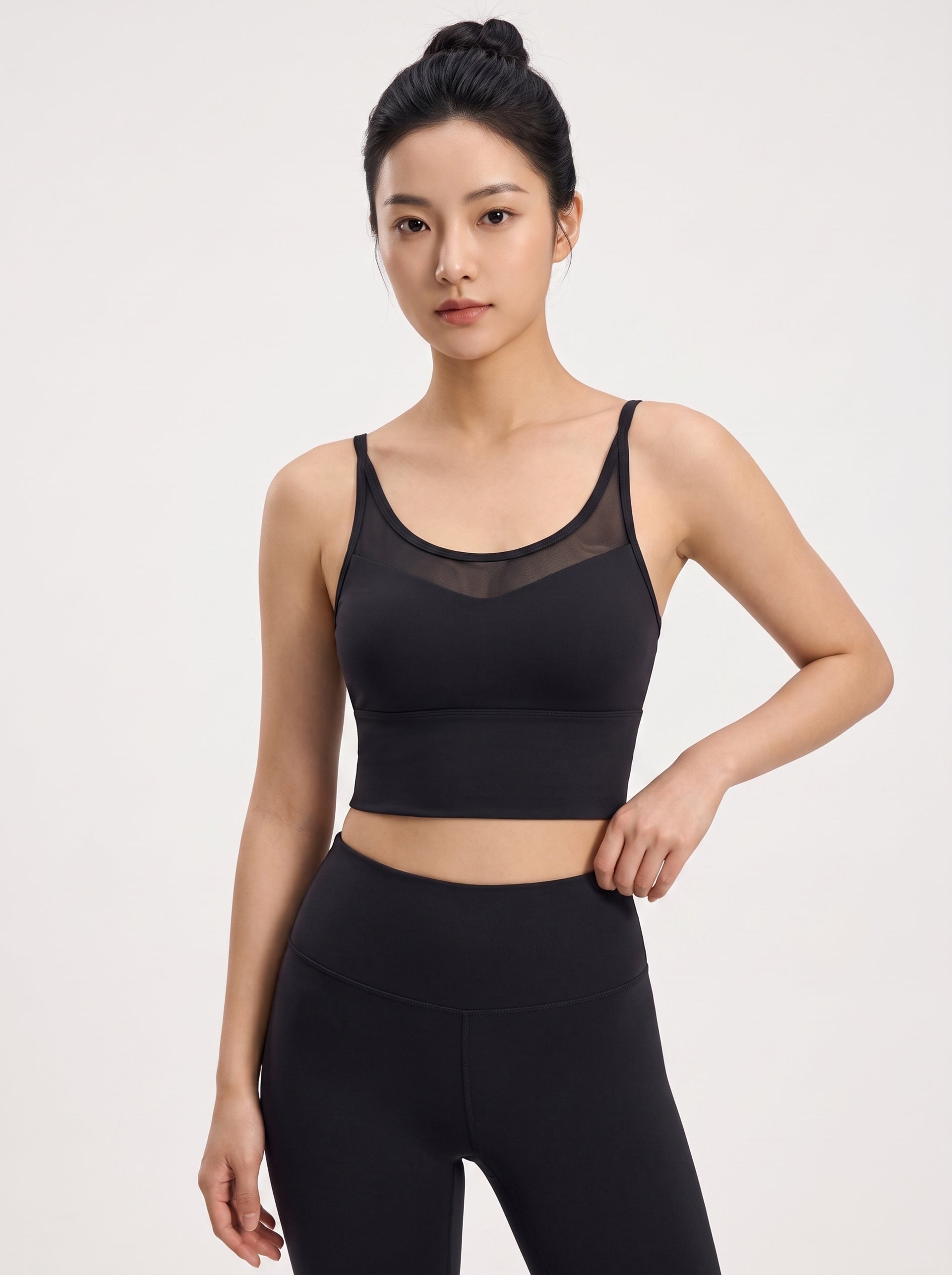 A woman modeling a black sports bra with a sheer mesh V-neckline and matching black high-waisted leggings against a light gray studio background.
