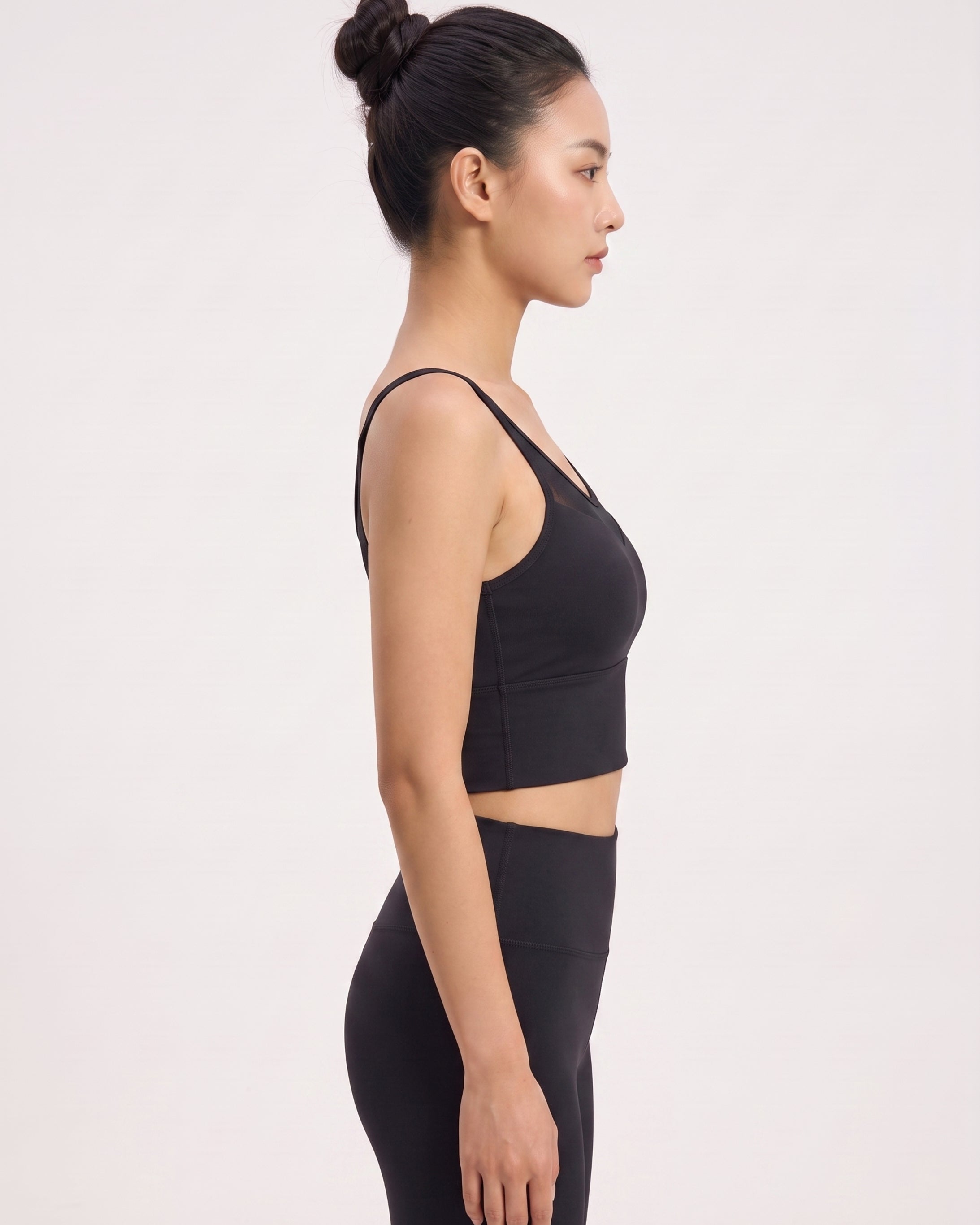 A woman in side profile wearing a black sports bra and matching black leggings against a plain light gray studio background.