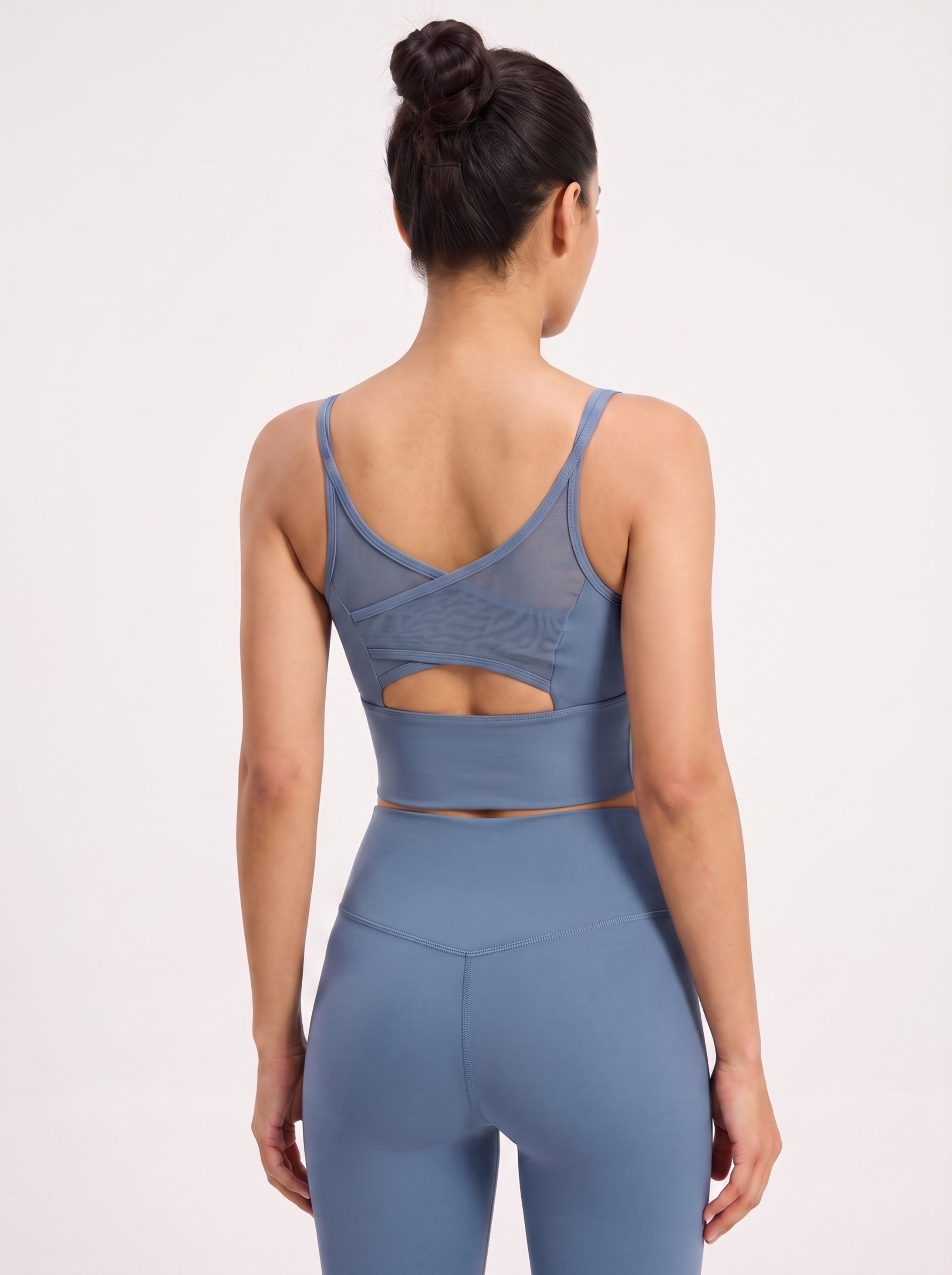 A woman shown from the back wearing a dusty blue athletic set, featuring a sports bra with a crossover back and cutout detail, paired with high-waisted leggings, against a plain white studio background.