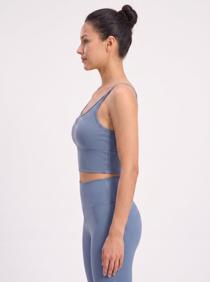 A woman in side profile wearing a matching slate blue activewear set, featuring a sports bra and high-waisted leggings, against a plain white studio background.