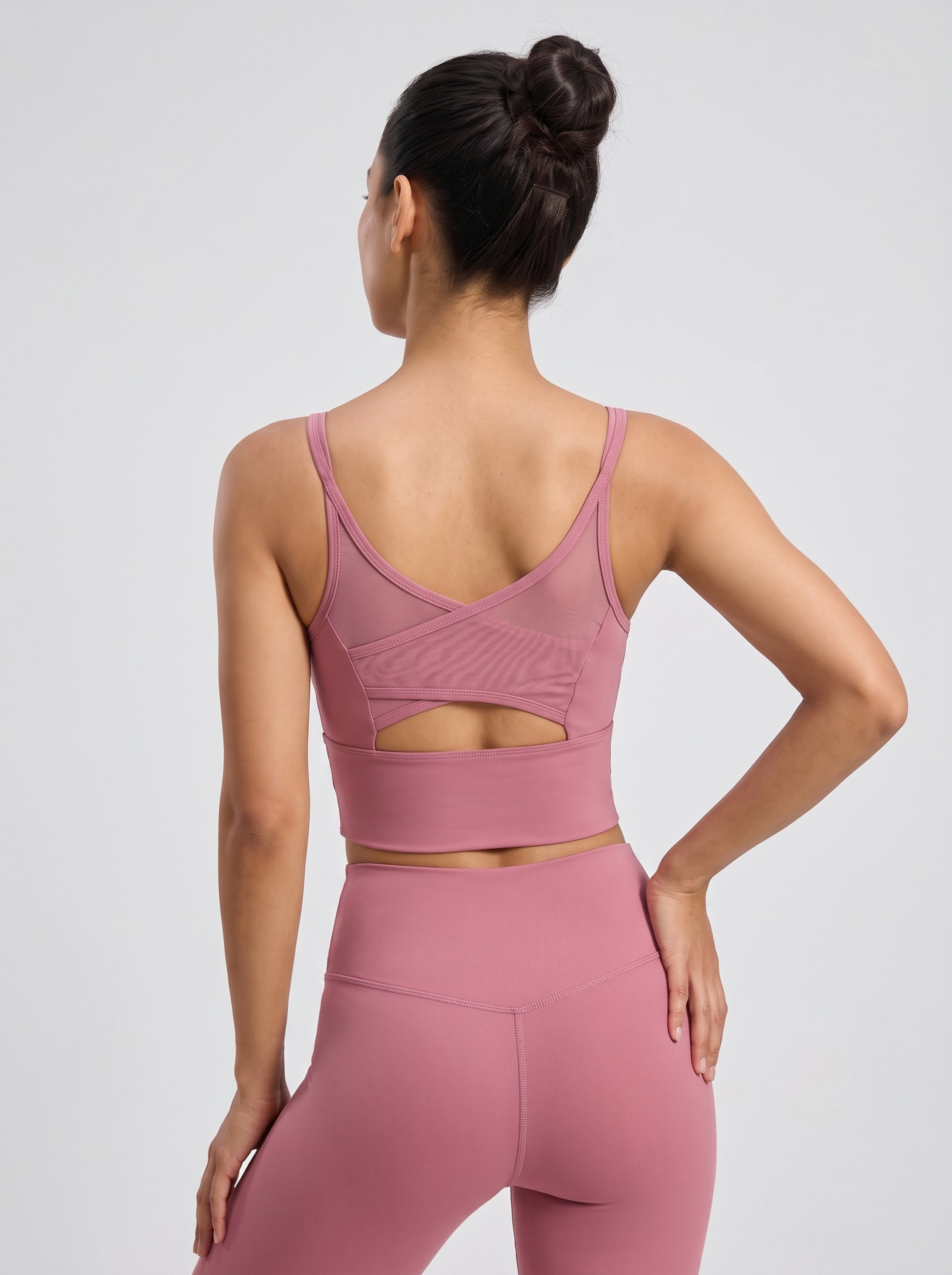 A woman shown from the back wearing a dusty pink athletic set, featuring a sports bra with a crossover criss-cross back design and a keyhole cutout, paired with matching high-waisted leggings, against a light gray studio background.