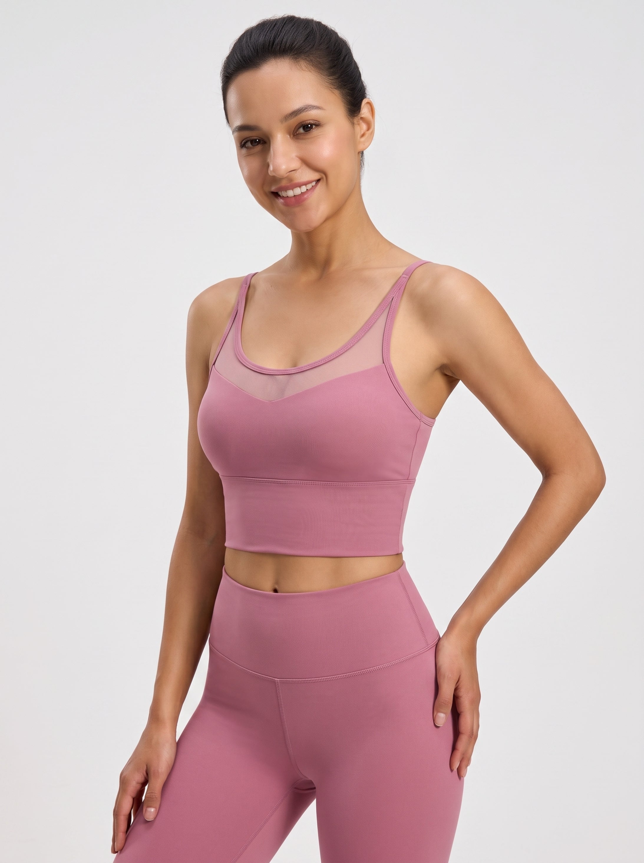 A woman modeling a dusty pink activewear set consisting of a sports bra with mesh detailing and matching high-waisted leggings, set against a plain light gray studio background.