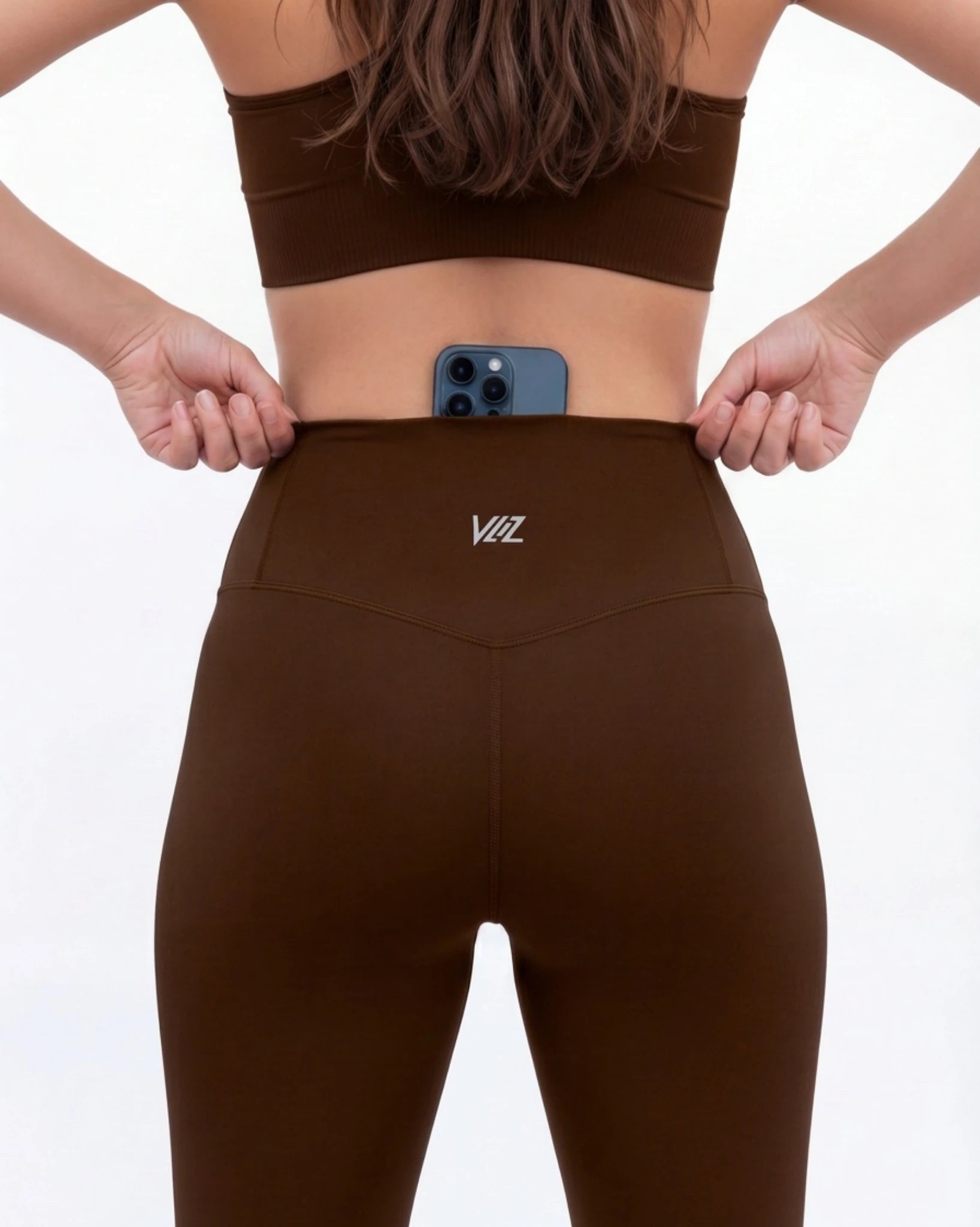 Person wearing brown leggings with a logo, holding a phone in front of their stomach on a white background